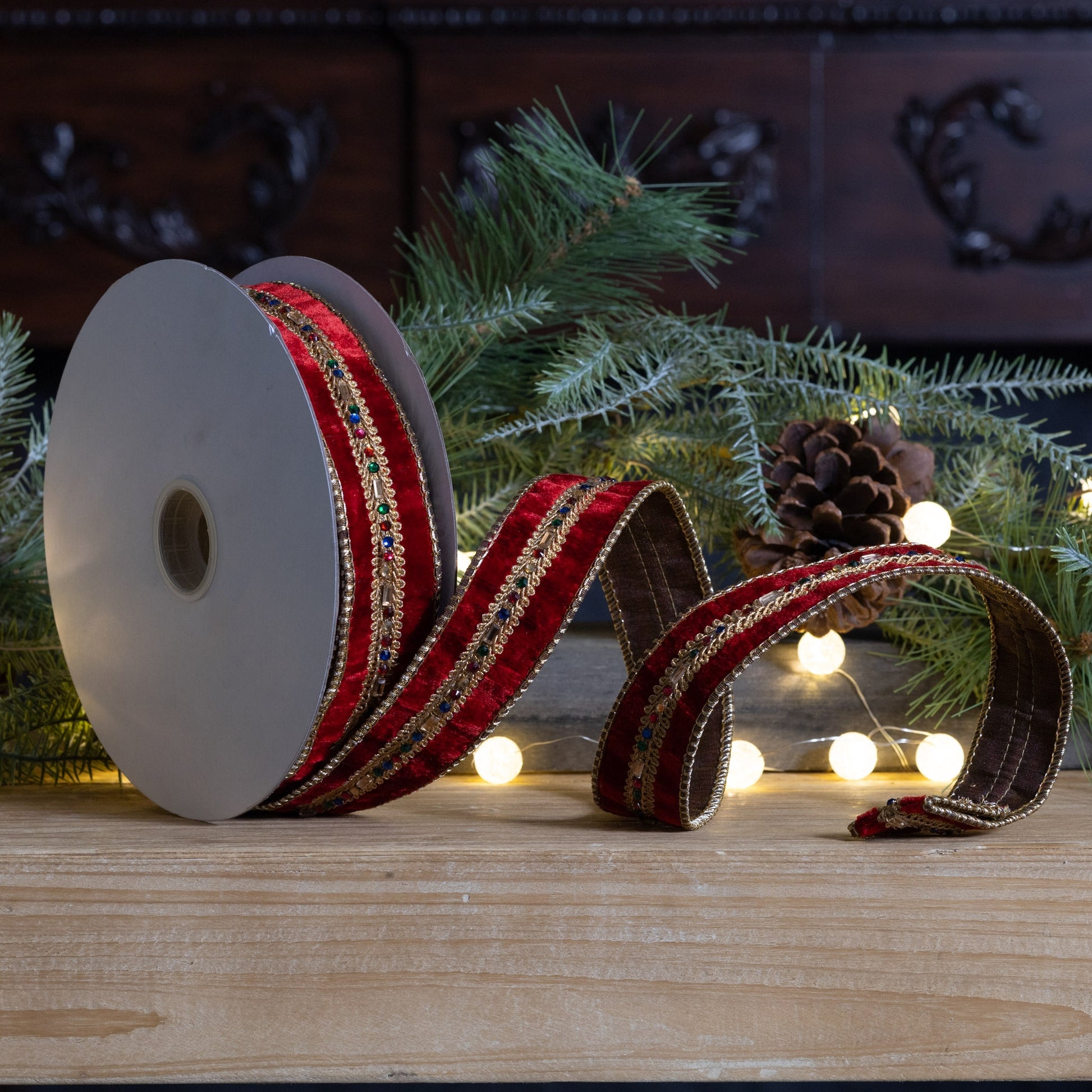 A roll of 1.5-inch wide red velvet ribbon with wired edges is partially unrolled on a wooden tabletop. The ribbon features an ornate gold embroidered center stripe accented with small multicolored jewel-like embellishments. Evergreen branches, pinecones, and warm white string lights create a festive Christmas background.